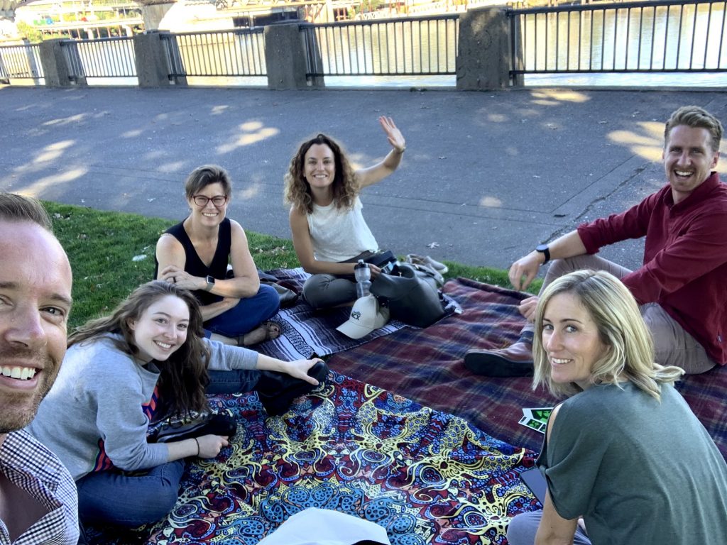 A group of NCX colleagues sit on blankets and talk in Tom McCall Waterfront Park in downtown Portland, Oregon