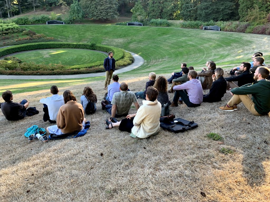 NCX colleagues sit on the green grass of Vietnam Veterans of Oregon Memorial in Washington Park, Portland, Oregon and listen to CEO Zack Parisa speak