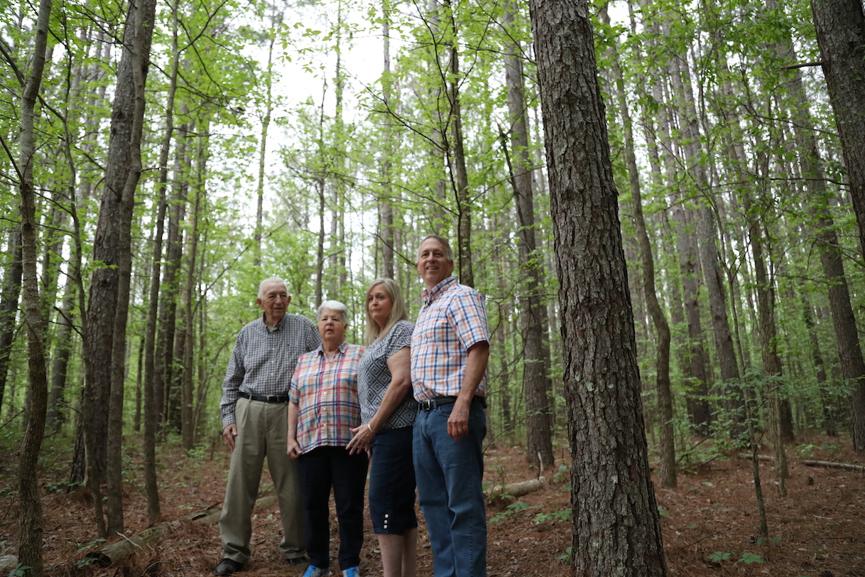 A multigenerational family of landowners stands together in a grove of trees on their land