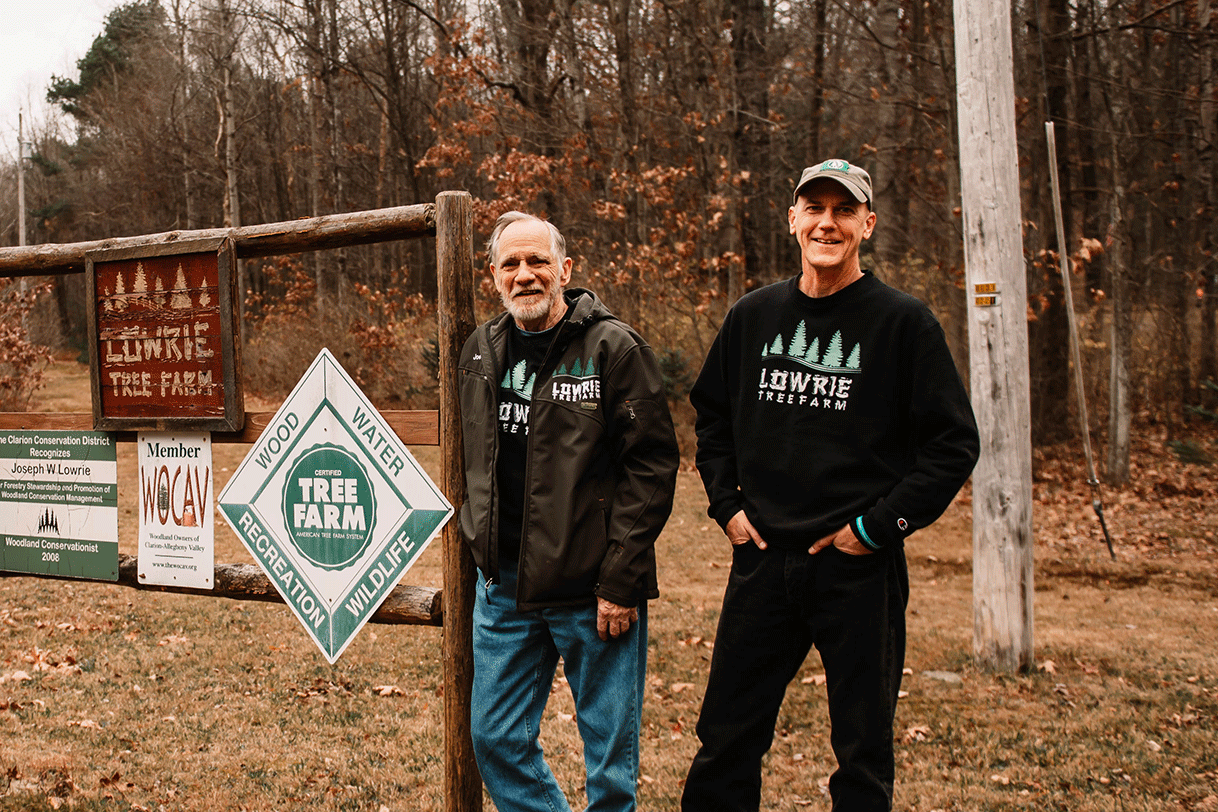 Father and son stand together in front of the Lowrie Tree Farm sign with their Pennsylvania forest in the background